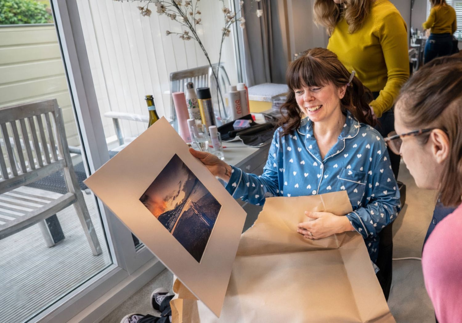 Woman smiling while unwrapping a framed photo print as a gift, with friends gathered around indoors.