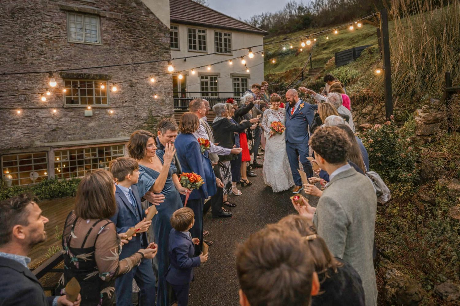 Wedding guests line up outdoors at dusk, string lights glowing, as bride and groom walk through a celebratory receiving line.