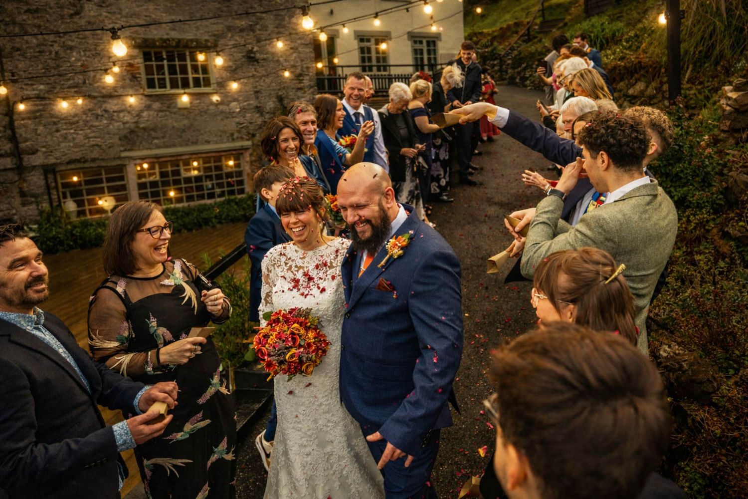 Bride and groom walk through cheering guests outdoors at rustic wedding venue with string lights.