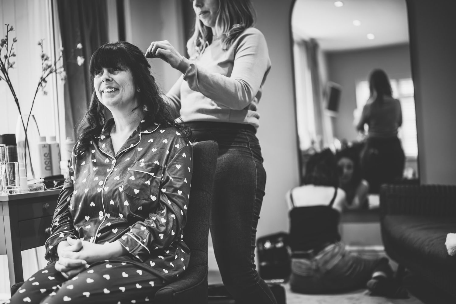 Black and white photo of a woman smiling as a hairstylist works on her hair in a bright salon setting.