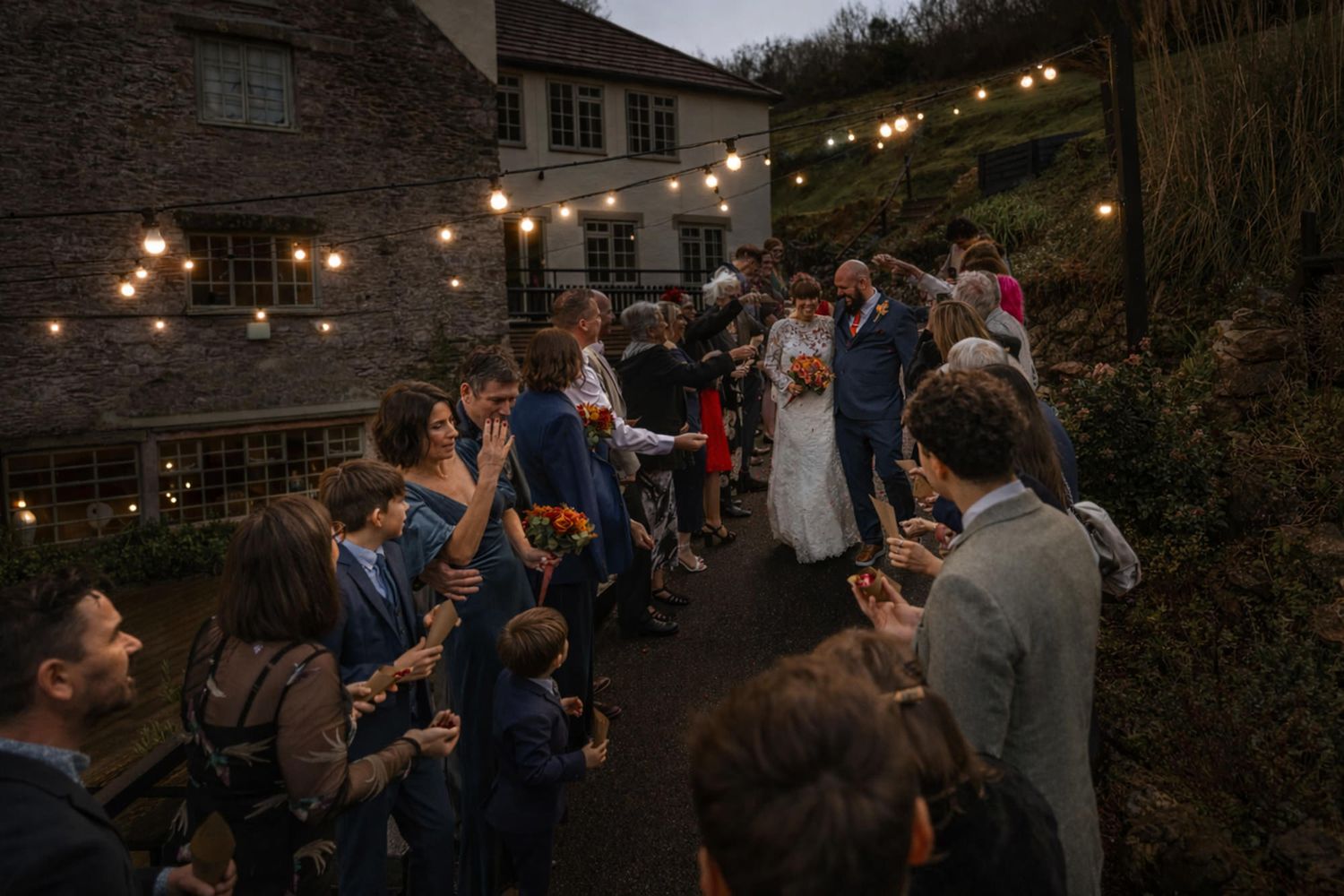 Wedding guests line a path under string lights as bride and groom walk through at dusk near a stone venue.