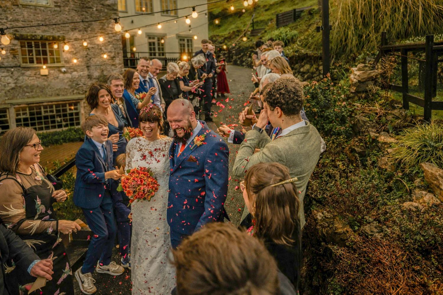 Bride and groom walk through confetti shower surrounded by wedding guests outside a rustic stone venue.