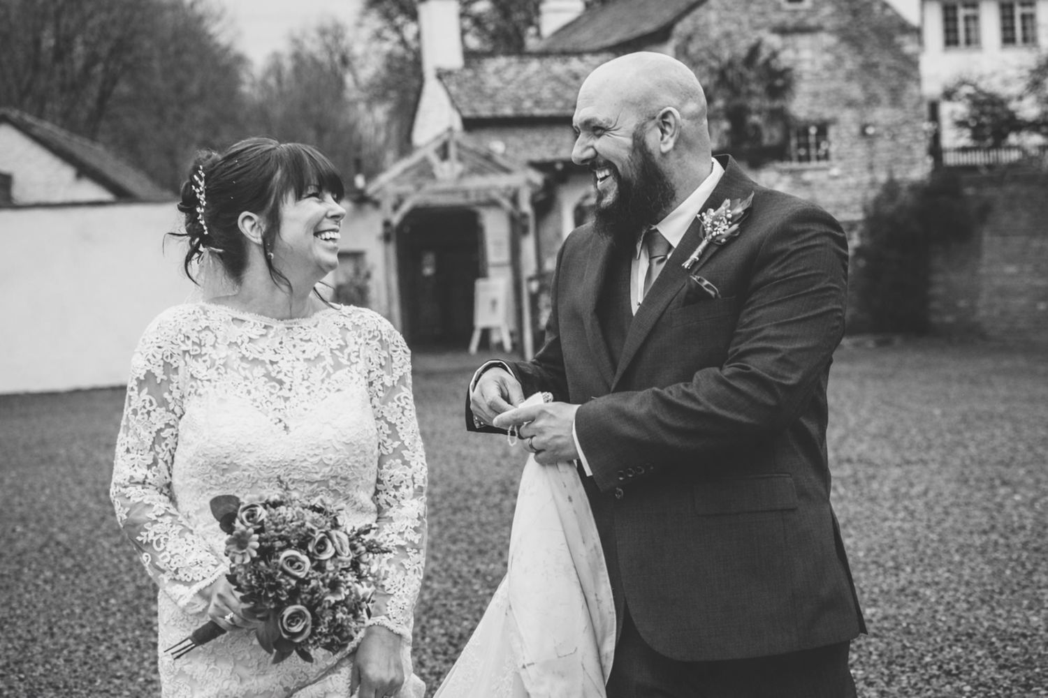 Black and white wedding photo of smiling bride in lace gown and groom in suit holding hands outdoors.