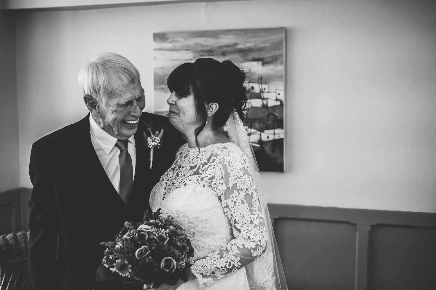 Black and white photo of a bride in lace gown holding bouquet, sharing a tender moment with an elderly man in a suit.
