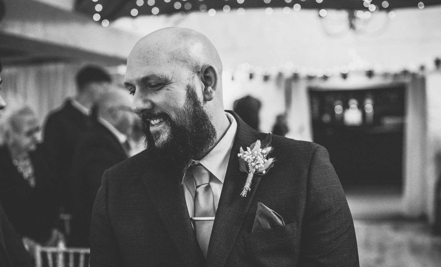 Smiling bald groom with beard wearing suit and boutonniere at wedding reception in black and white photo.