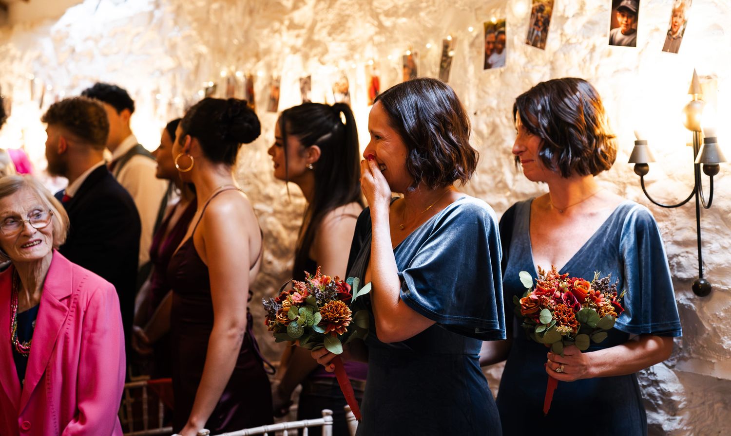 Bridesmaids in blue velvet dresses holding autumn bouquets laugh together in a rustic stone wedding venue.