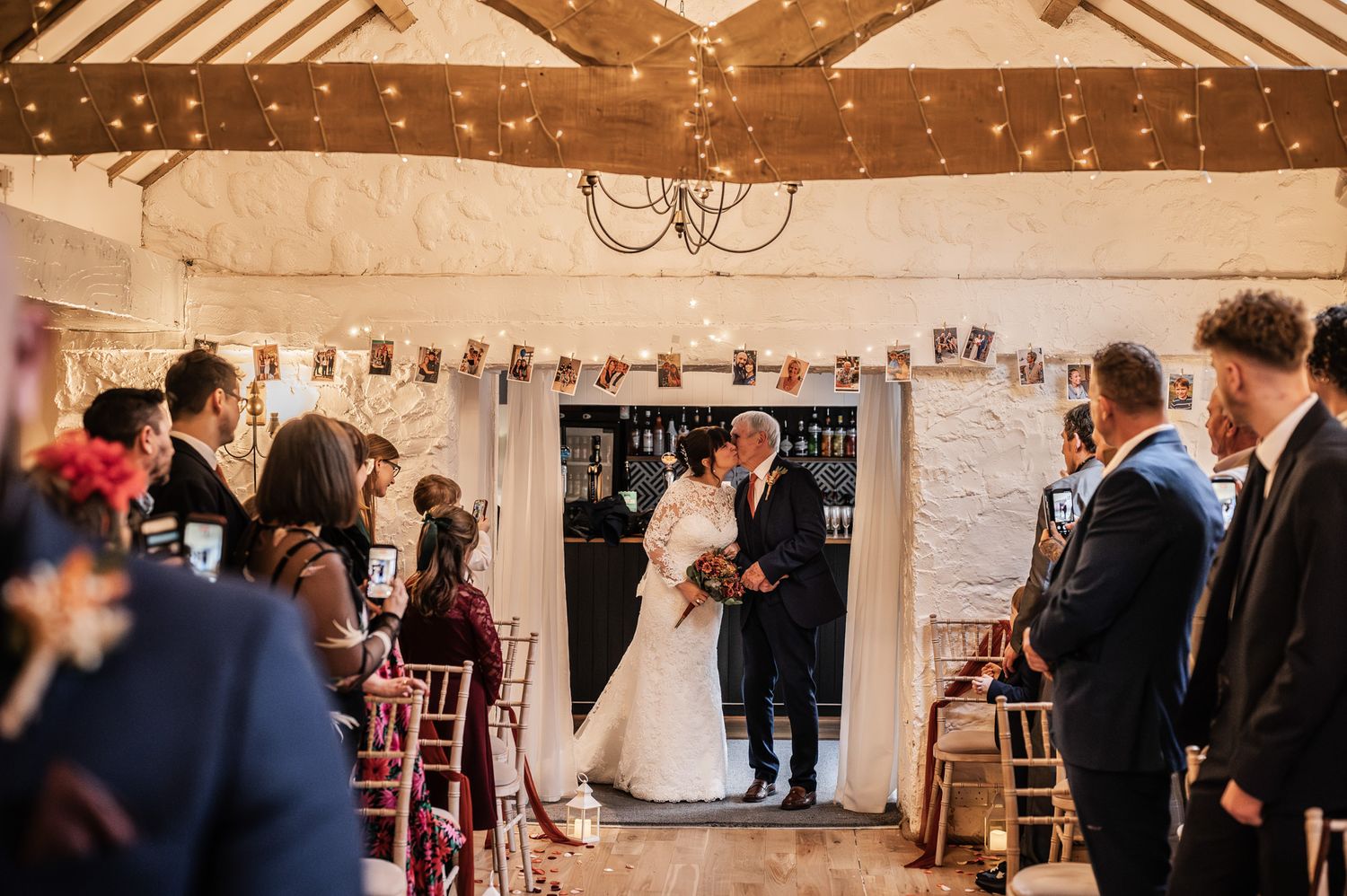 Bride and groom share a kiss at rustic barn wedding ceremony with fairy lights and guests seated in gold chiavari chairs.