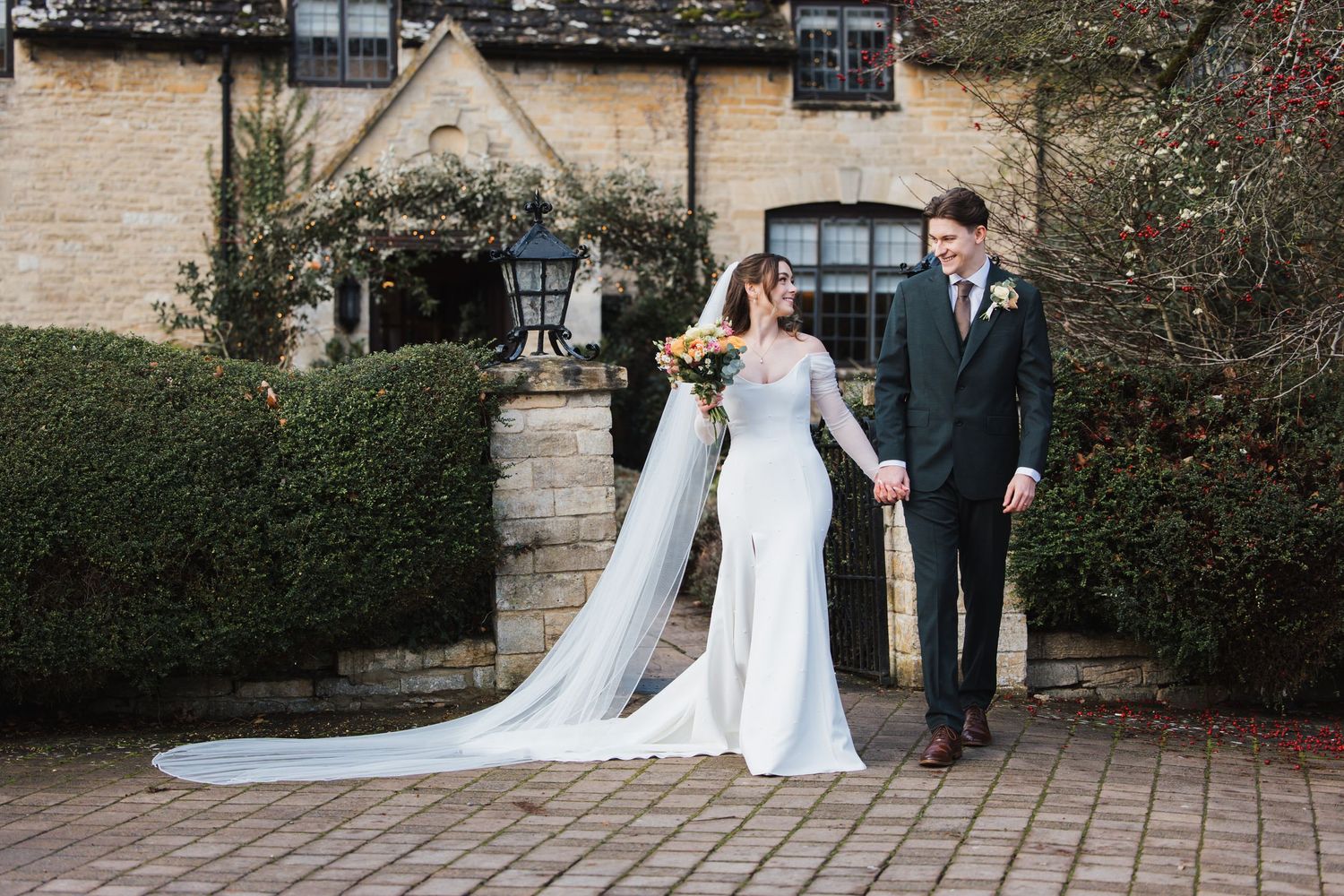 Bride and groom walking hand in hand outside a stone manor house, bride wearing a long veil and groom in a dark suit.