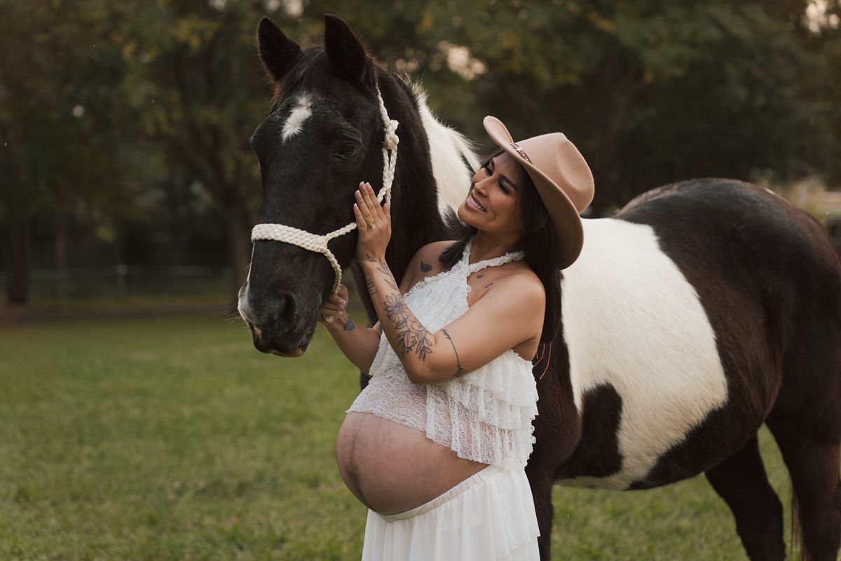 Pregnant woman in white dress and tan hat touching a black and white paint horse in a green outdoor setting. Maternity photoshoot Sydney