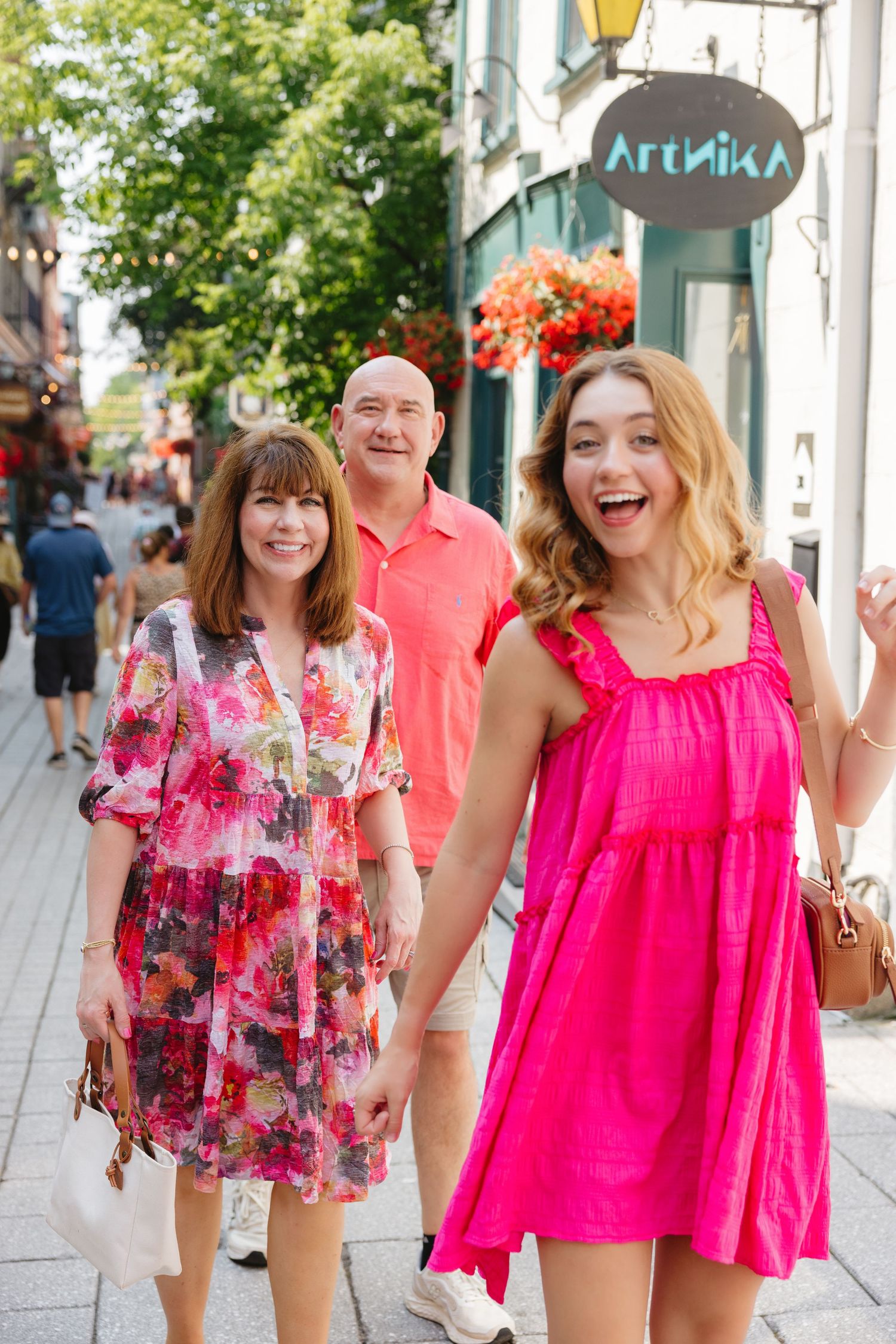 A joyful family of three walks along a vibrant outdoor shopping street, wearing bright pink and floral summer outfits.