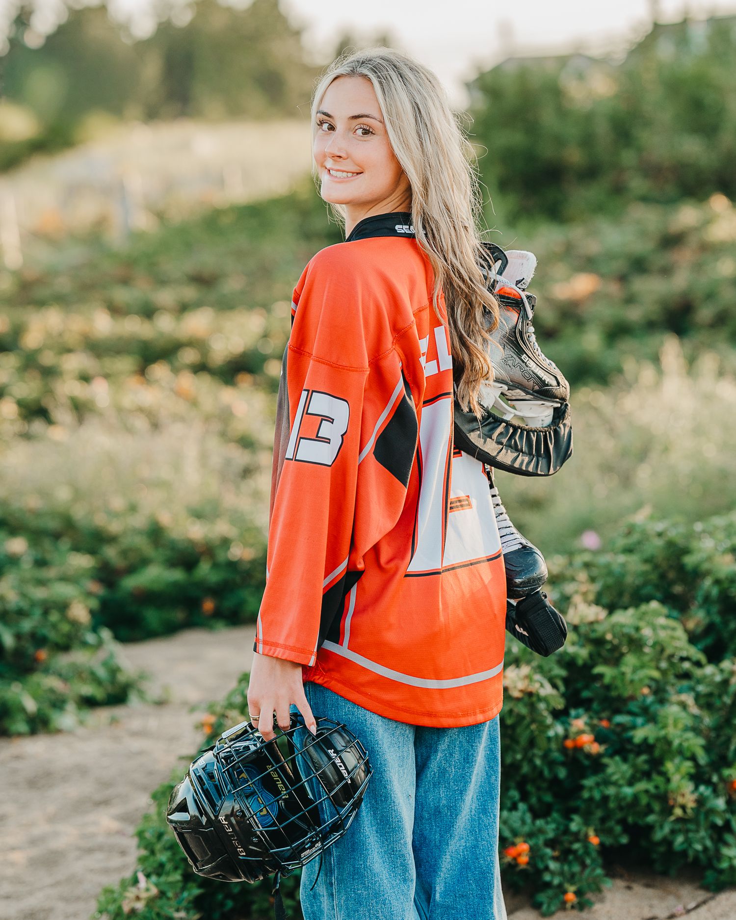 high school senior in red hockey jersey holding helmet and gloves, posing outdoors in a flower garden.