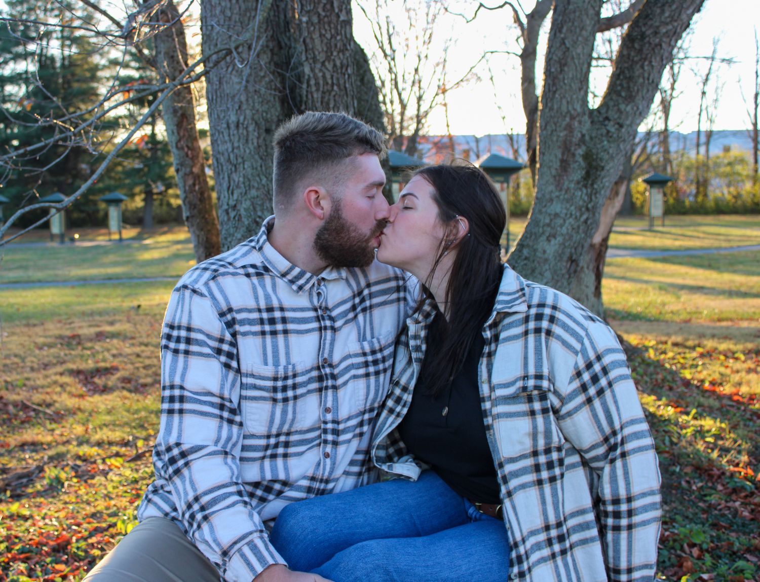 Couple sharing a kiss outdoors, sitting on grass near trees in matching plaid flannel shirts on a sunny day.