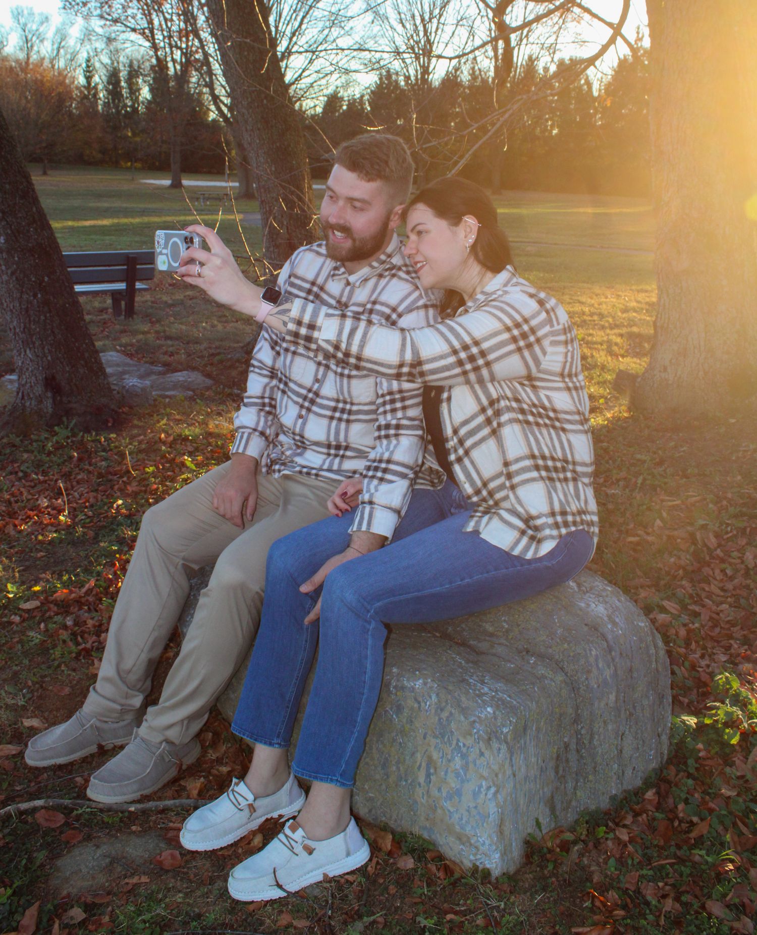 Couple in matching plaid shirts taking a selfie on a rock in a sunny park during golden hour.
