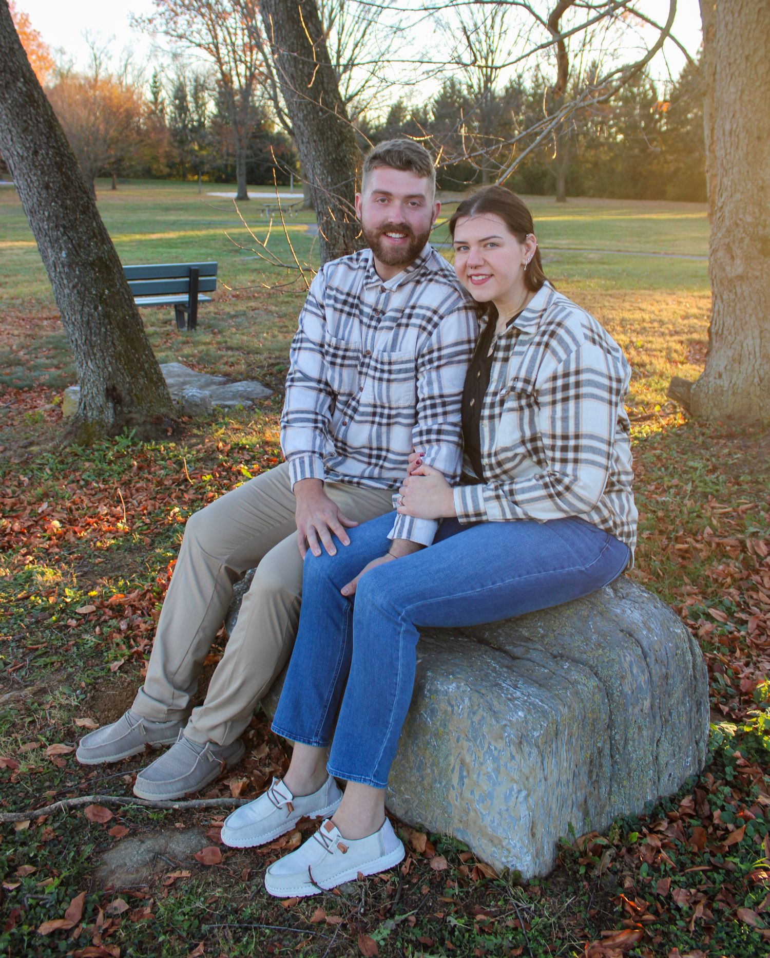 Couple in matching plaid shirts sitting on a stone in a park with autumn leaves on the ground.