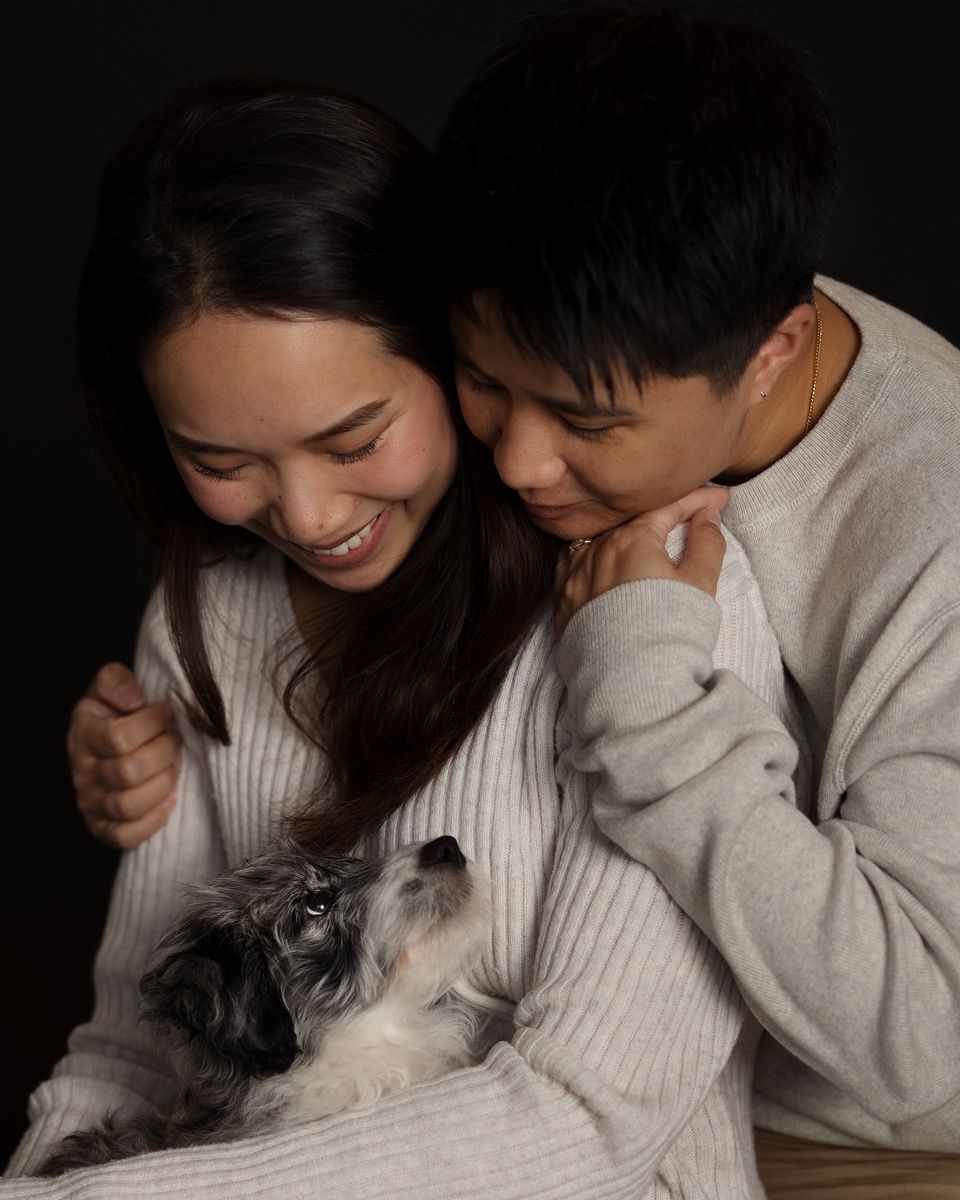 Smiling couple in matching cream sweaters cuddling with a small broodle dog against a dark background in rouse hill photoshoot