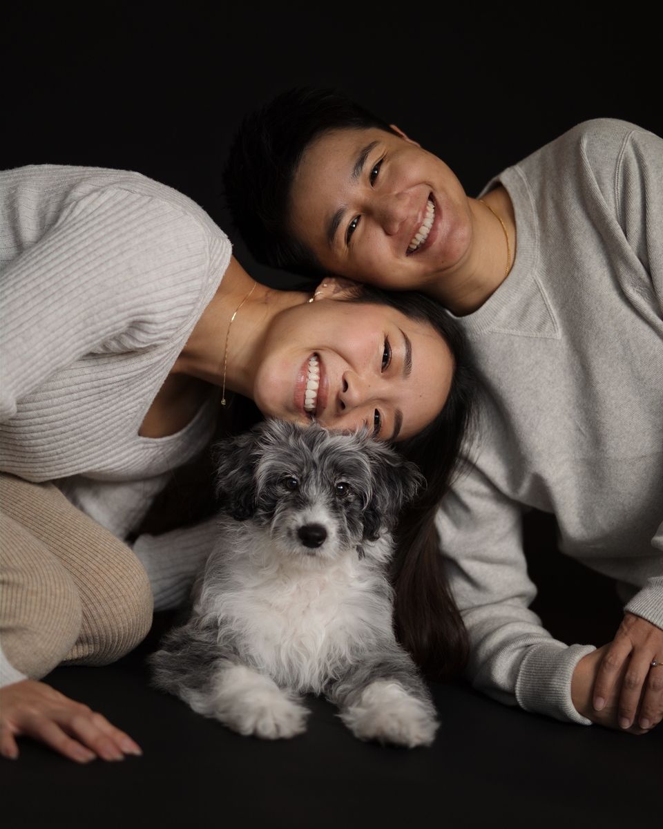 Two smiling women pose with a fluffy gray and white puppy against a dark background in rouse hill photoshoot