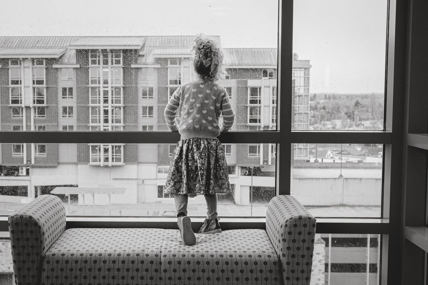 Black and white photo of a young girl standing on a chair, gazing out a large window at city buildings.