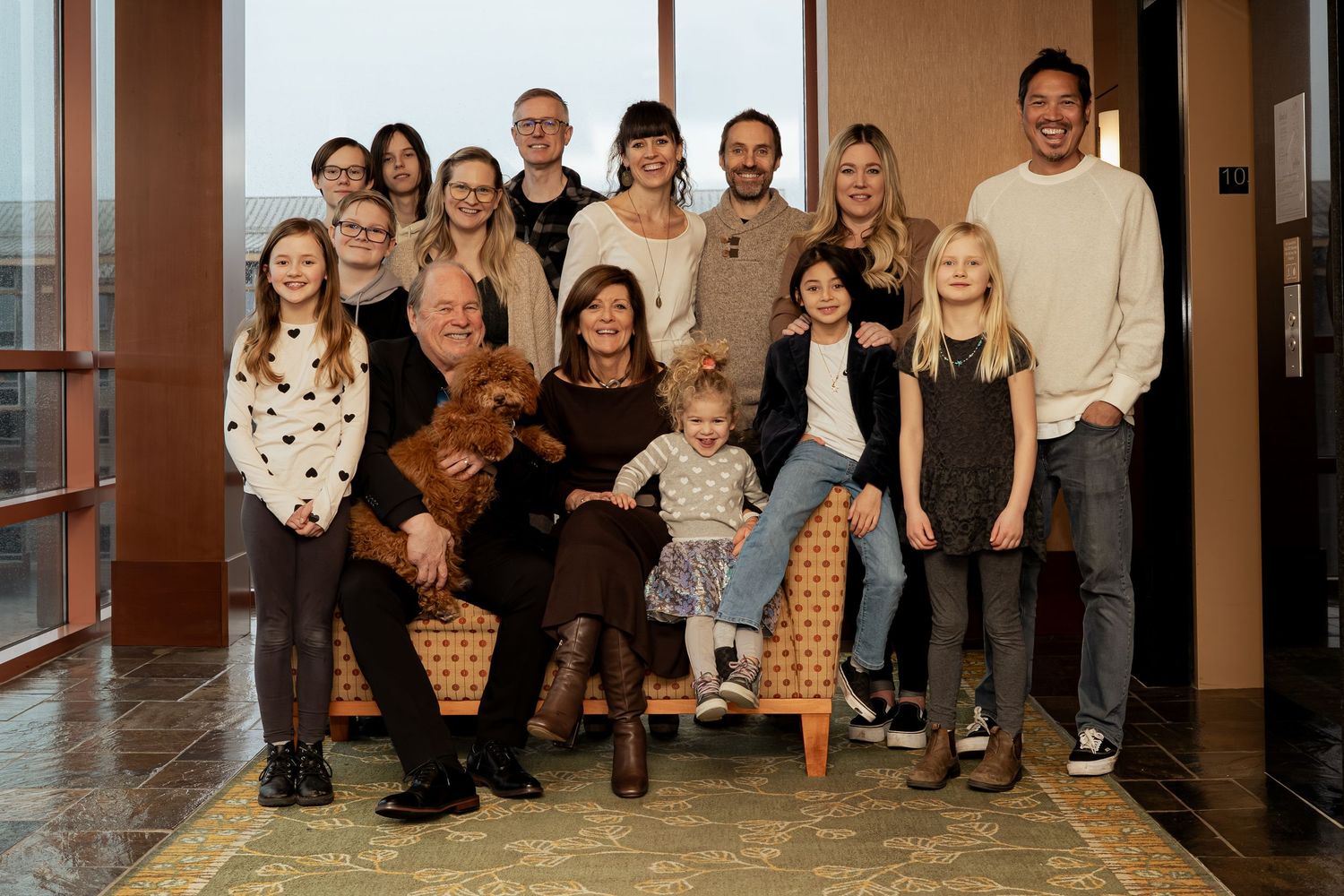 Large multigenerational family group portrait smiling together indoors, including children, adults, and a brown dog.