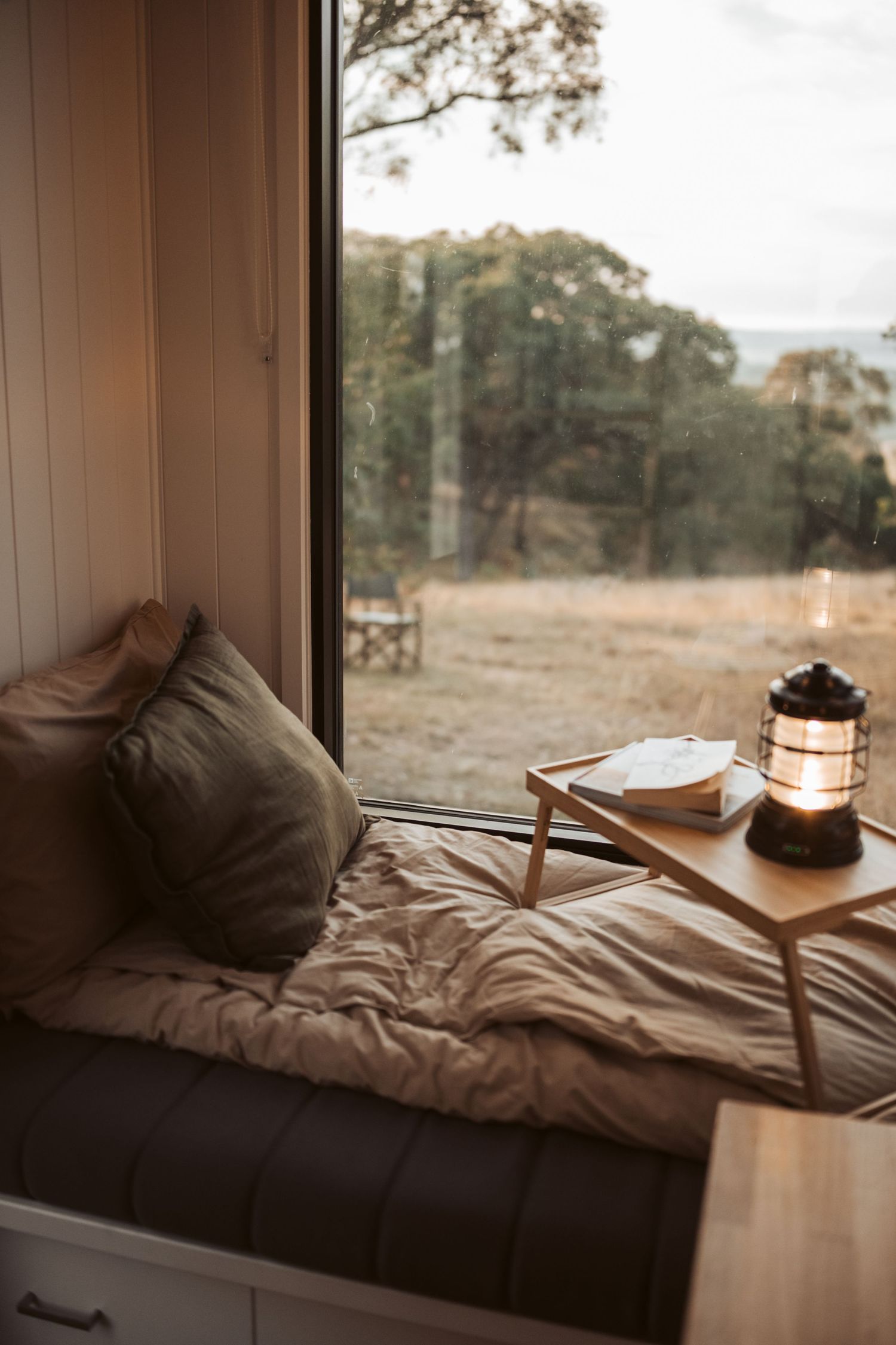 Cosy window nook with cushioned bench, wooden breakfast tray and lantern overlooking Australian bush with distant eucalyptus trees.