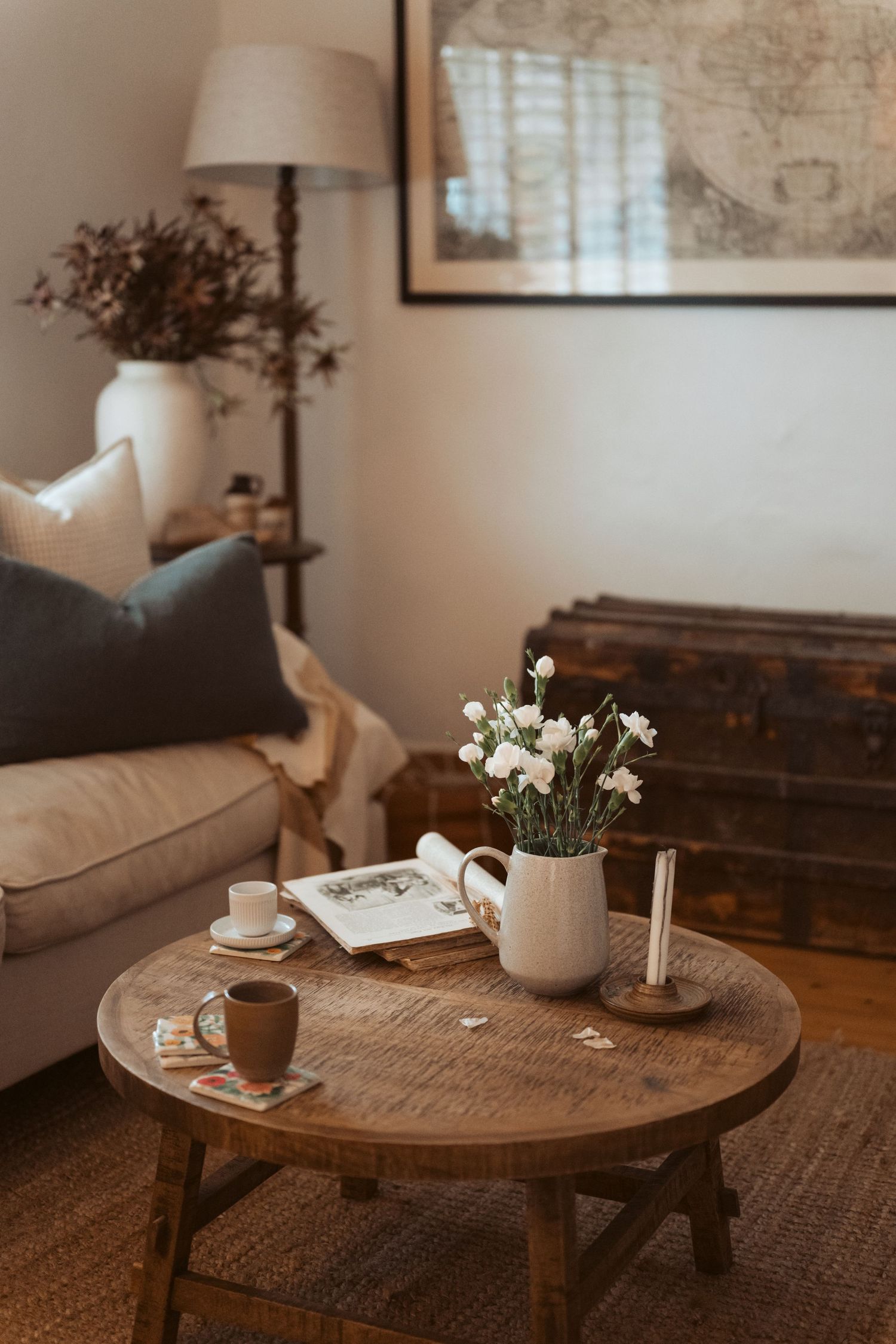 Rustic wooden coffee table with white flowers, teacup and magazine. Vintage trunk and neutral sofa create relaxed country cottage feel.
