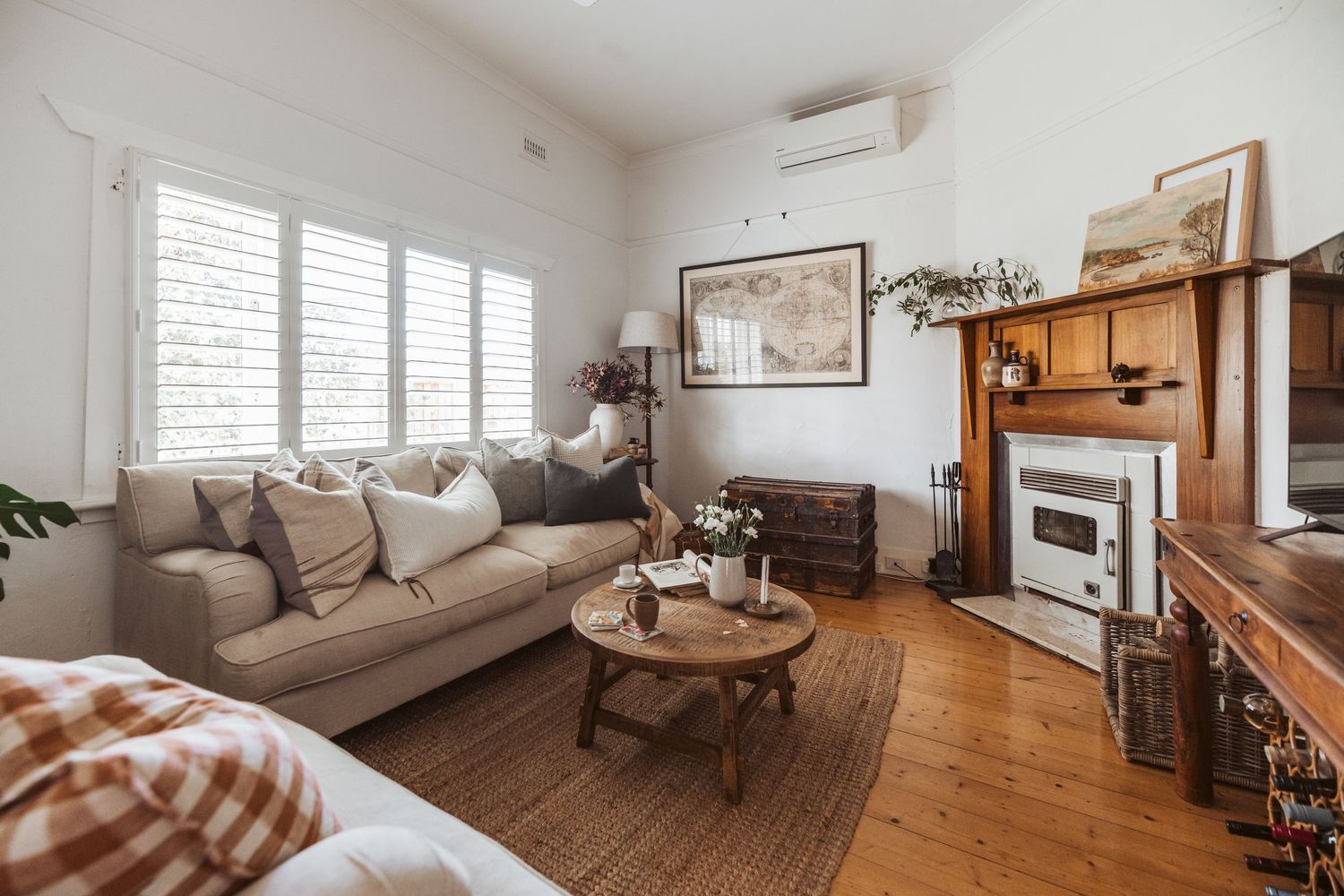 Bright coastal living room with plantation shutters, linen sofa, wooden coffee table and jute rug surrounding vintage fireplace.