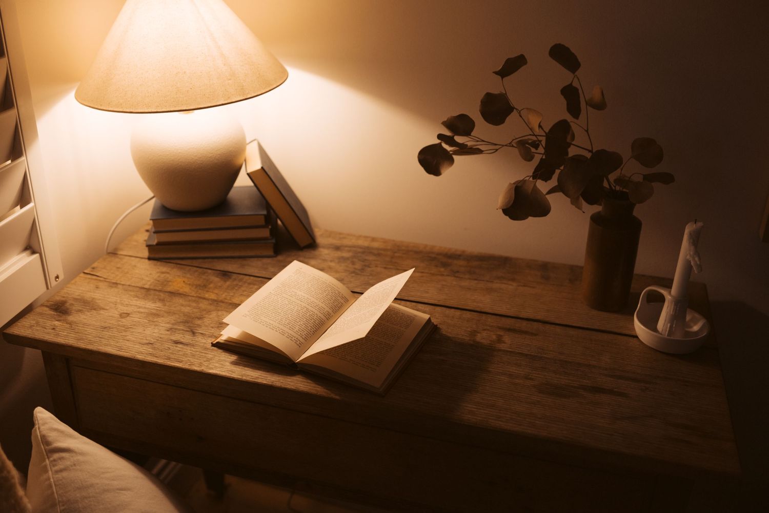 Intimate bedside scene with open book under warm lamplight. Wooden table, dried eucalyptus in vase creating serene reading nook.