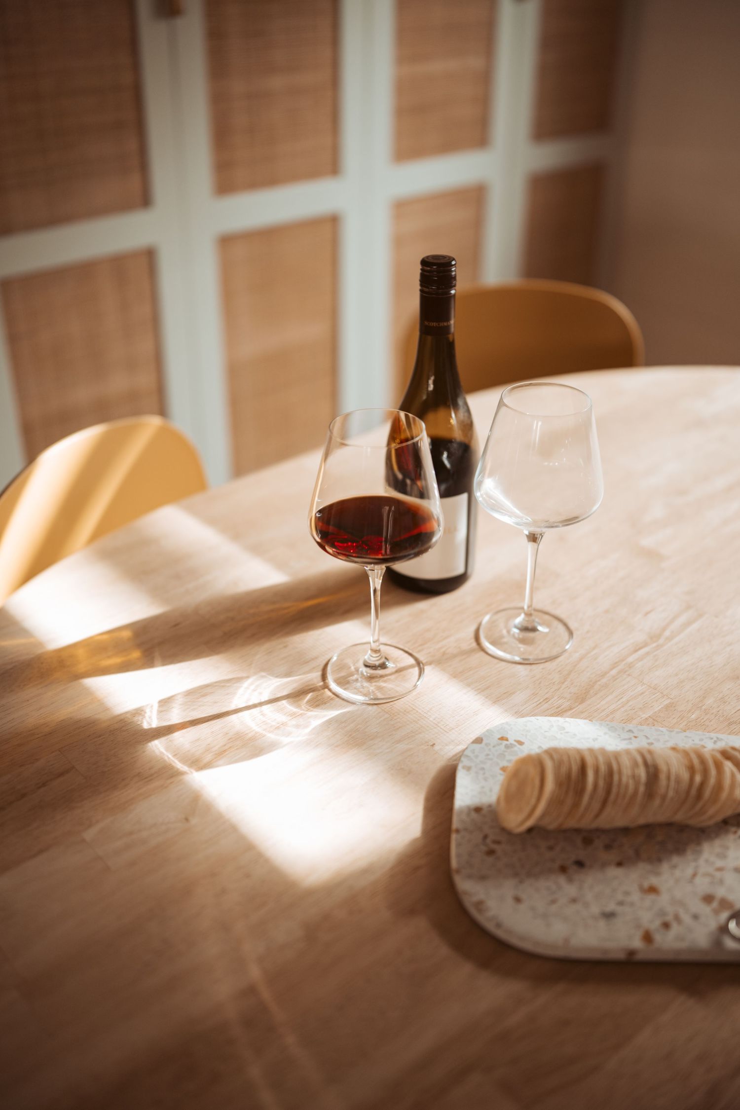 Sunlit wooden table with red wine, glasses and marble cheese board. Warm light streams through window blinds in rustic setting.