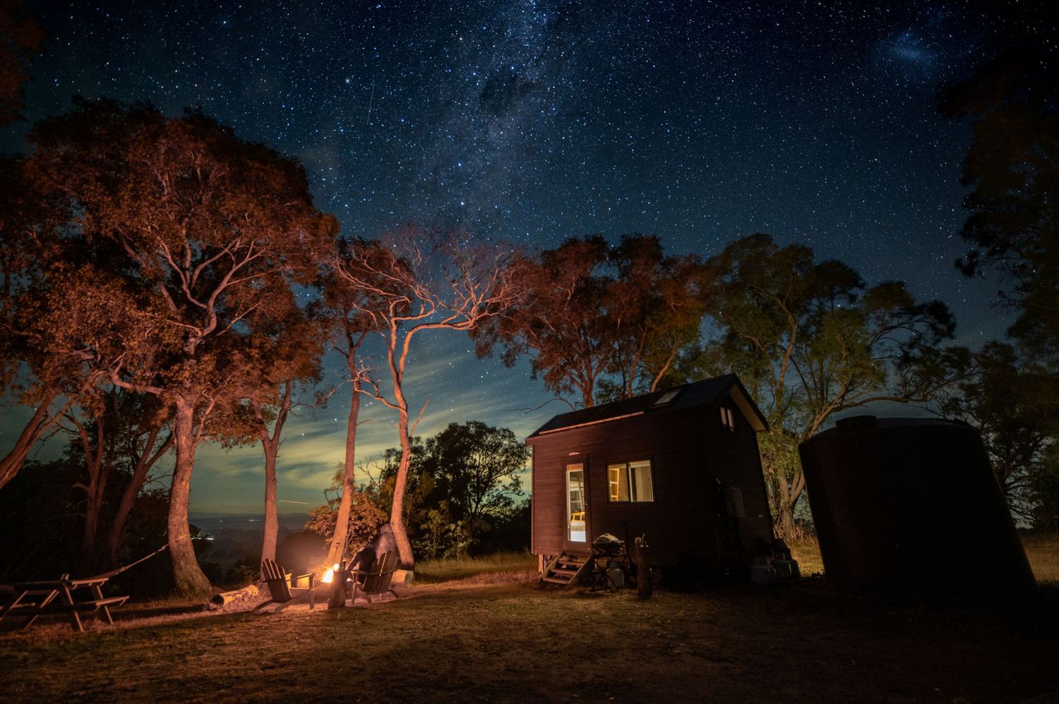 Cosy tiny cabin in Australian bush at night with campfire, illuminated by golden light under starry sky with visible Milky Way.