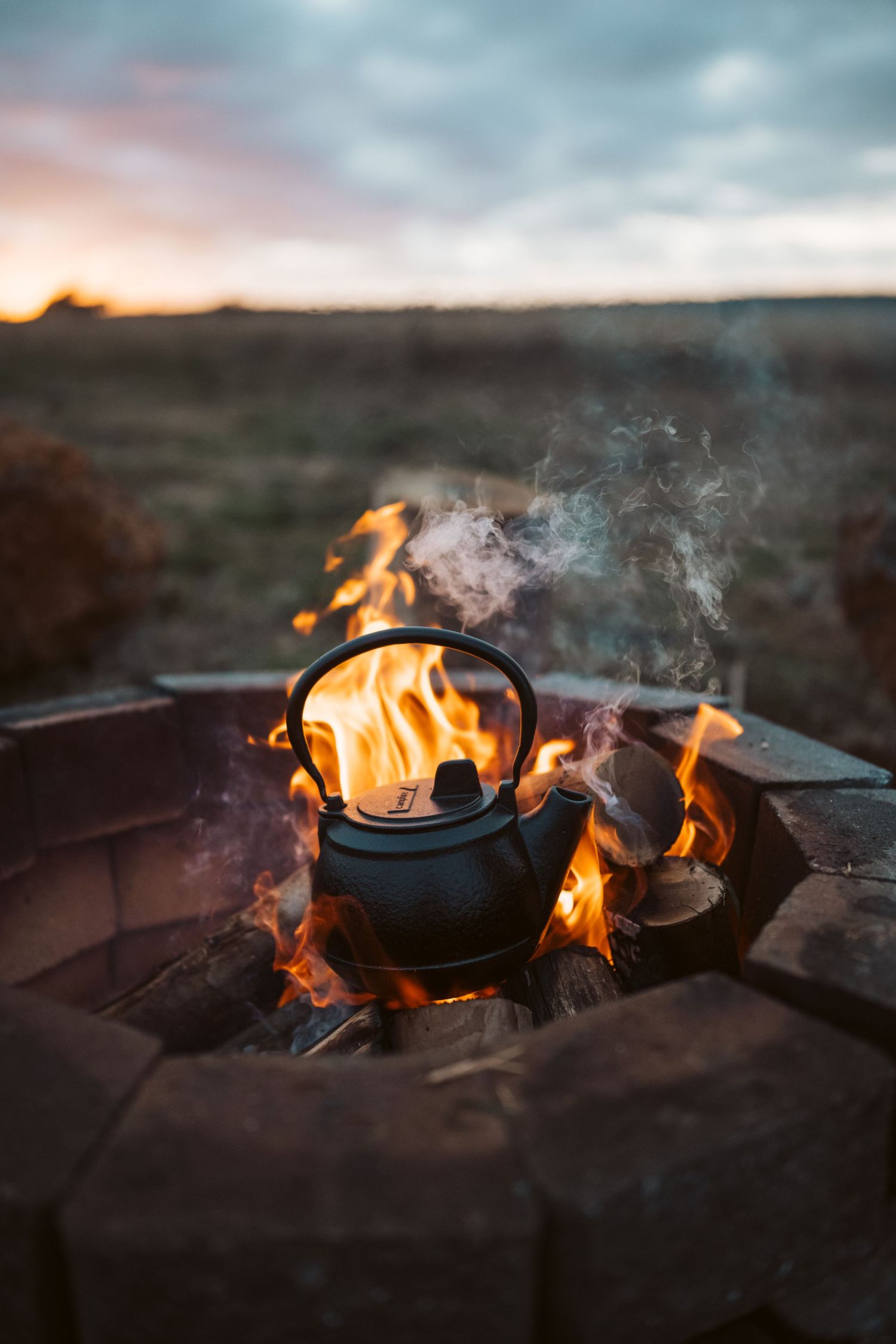 Cast iron kettle on open fire pit at sunset. Flames dance around black pot with smoky outback views beyond the stone surround.