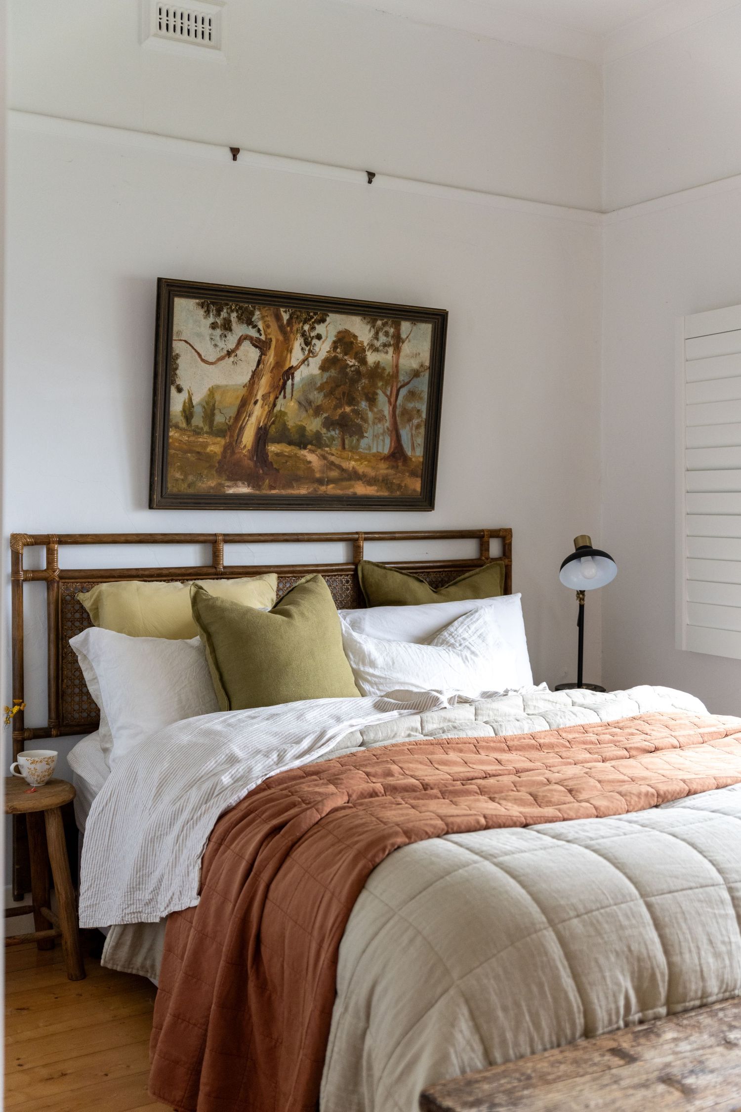 Cozy bedroom with bamboo headboard, white and terracotta bedding, sage green pillows. Framed landscape painting of gum trees hangs.