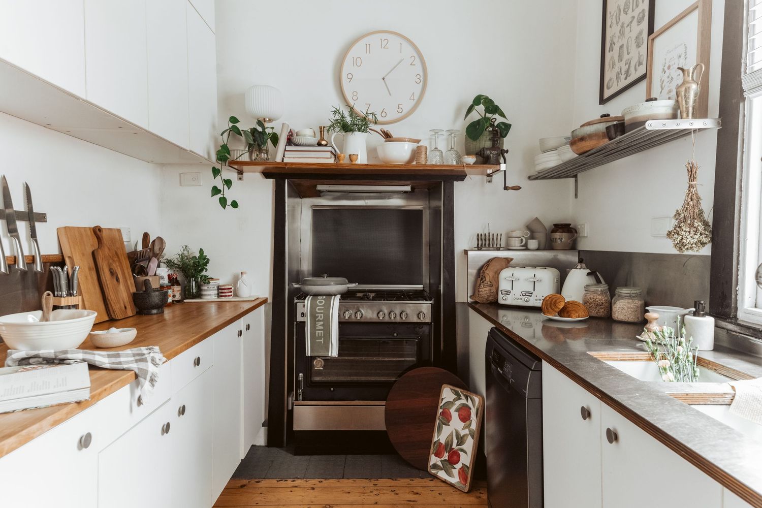 Compact farmhouse kitchen with white cabinetry, wooden countertops and vintage wood stove. Open shelving displays plants and kitchenware.