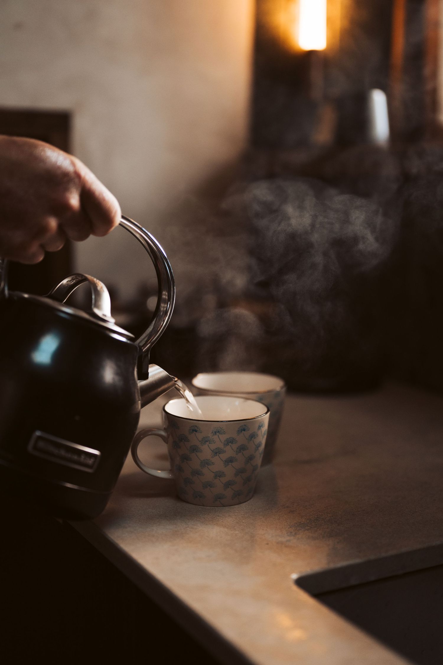Steaming kettle pouring hot water into patterned cups, creating warm morning atmosphere in cosy rustic cabin kitchen.