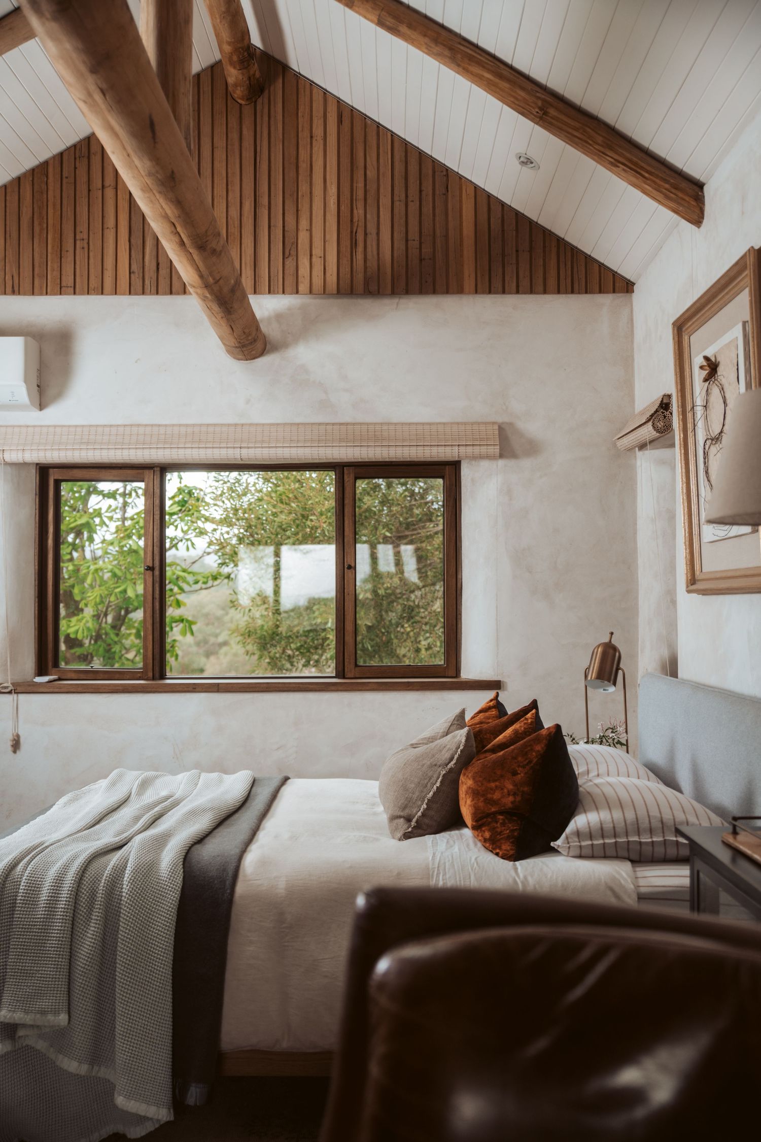 Bright, airy bedroom with rustic timber beams, white walls, large windows with bush views, and comfortable bed with neutral linens.