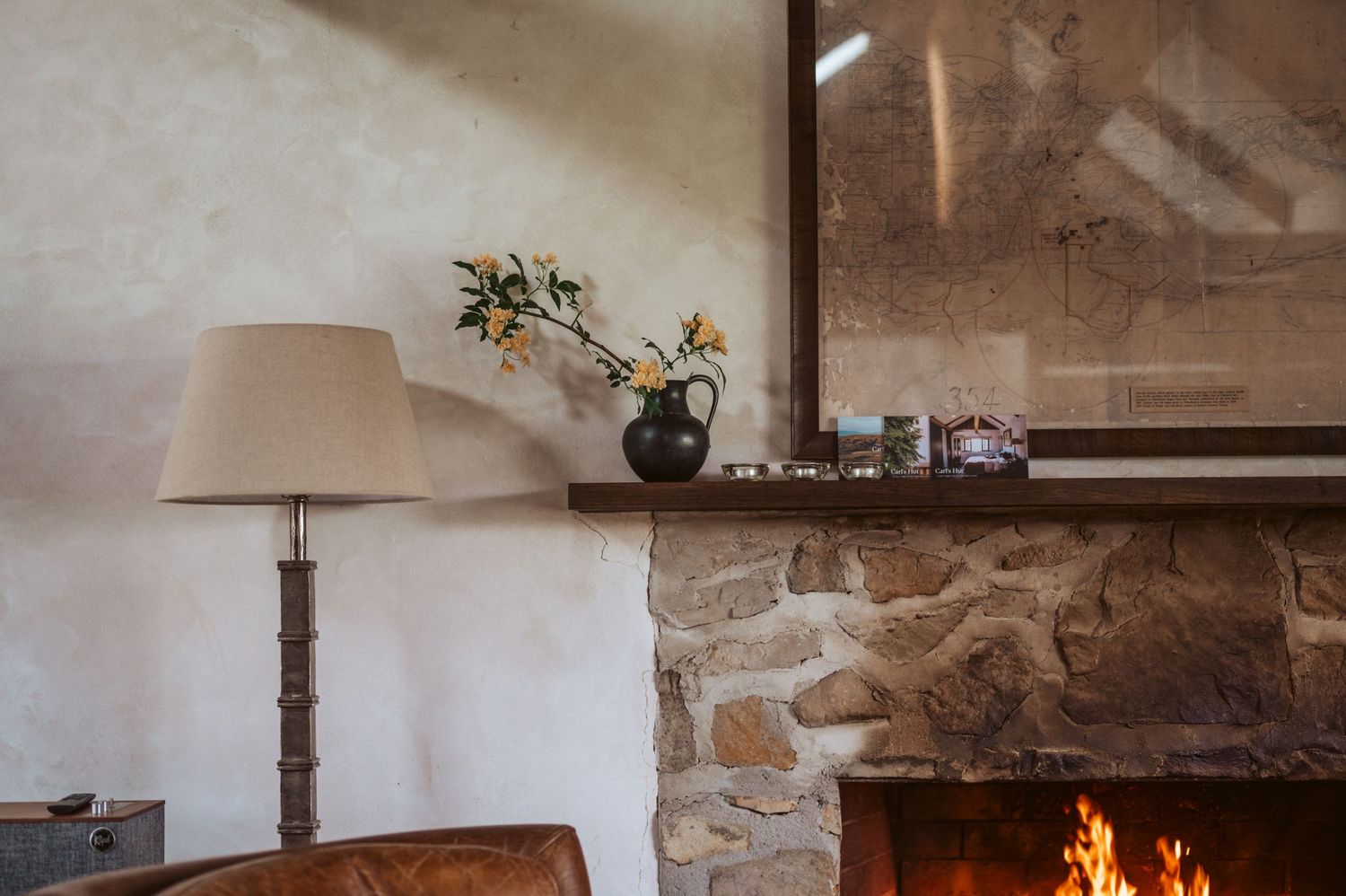 Cosy fireplace with stone surround, warm fire burning, featuring neutral décor with yellow flowers and wooden lamp in rustic setting.