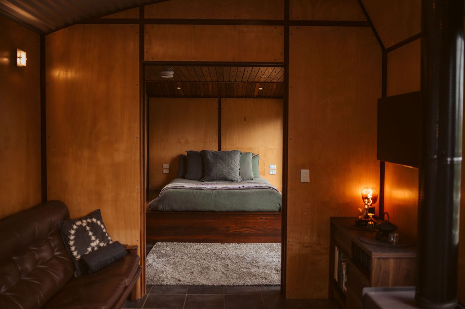 Rustic cabin interior with wooden walls, featuring a separate bedroom with light blue bedding visible through doorway.