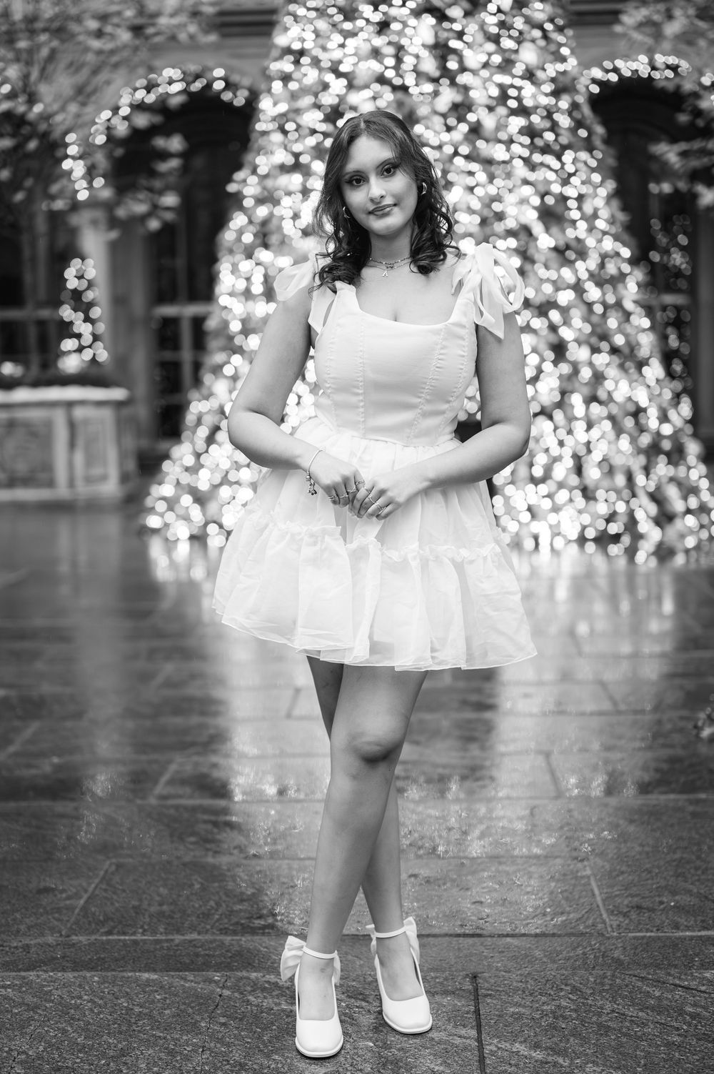 Woman in white ruffled dress standing before illuminated Christmas tree in black and white photo.