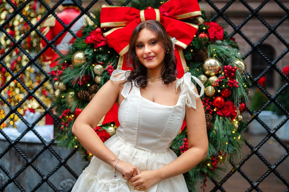 Young woman in white dress smiles in front of festive Christmas wreath with red bow outdoors.