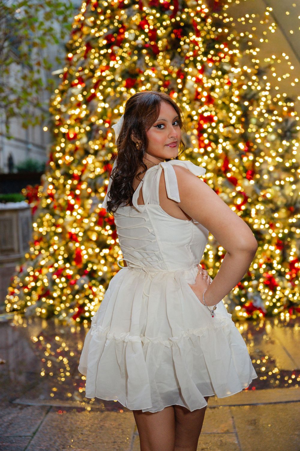 Woman in white dress posing in front of a decorated Christmas tree with gold lights and red ornaments.