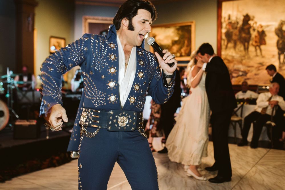 Elvis impersonator in blue sequin jumpsuit performing at a wedding reception while couple dances in background.