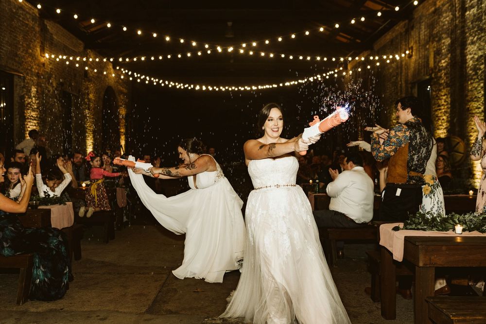 Two brides in white gowns celebrate at wedding reception with confetti poppers under string lights.