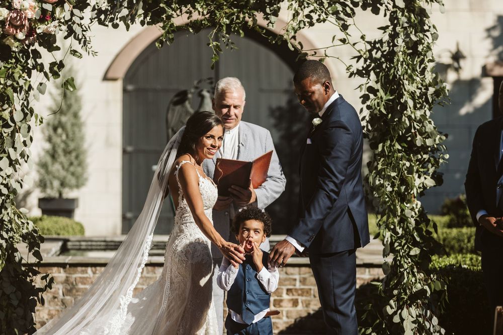 Bride and groom exchange vows under floral arch with young boy at outdoor wedding ceremony.