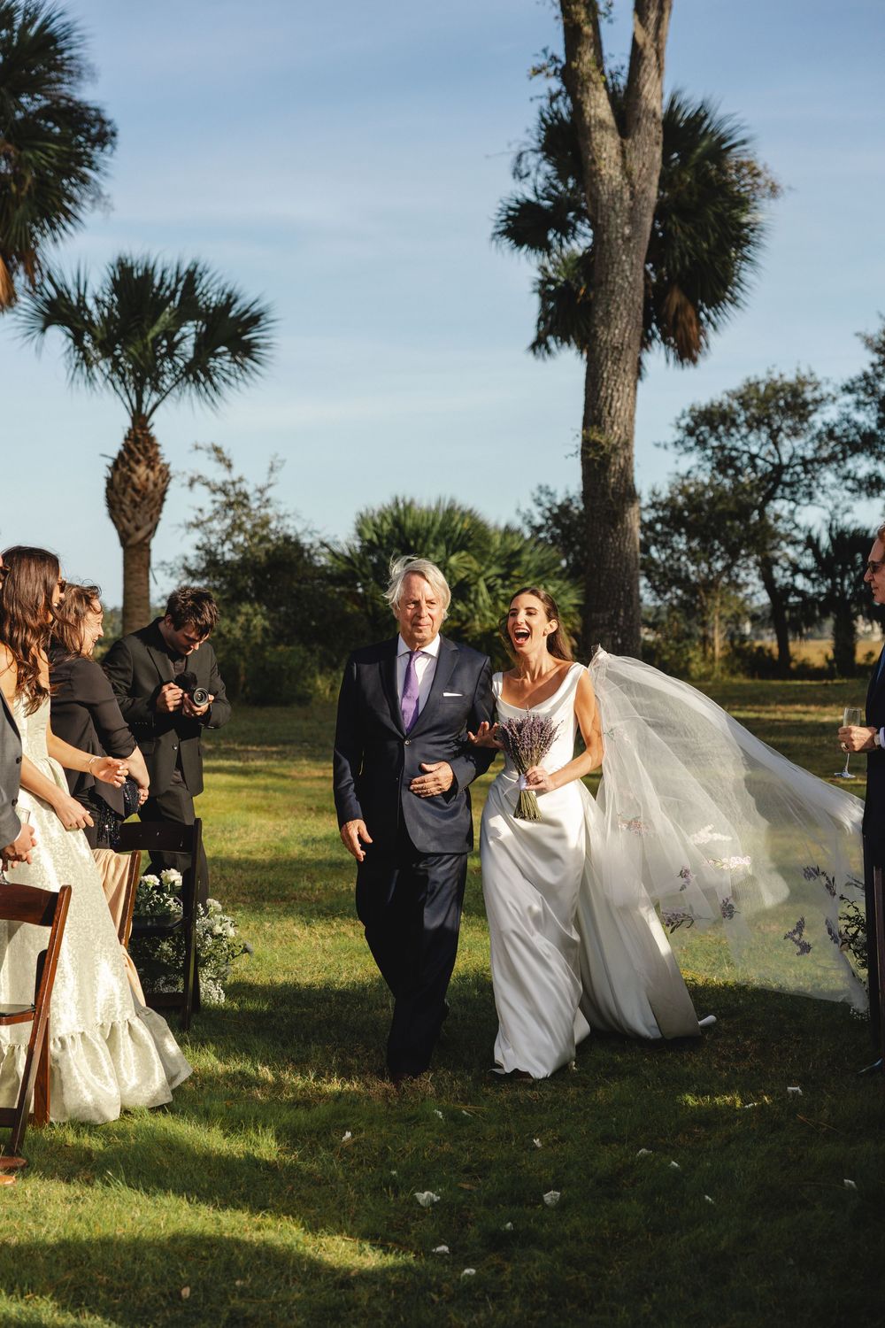 Bride laughing joyfully while walking down outdoor aisle with older man, palm trees and wedding guests in background.