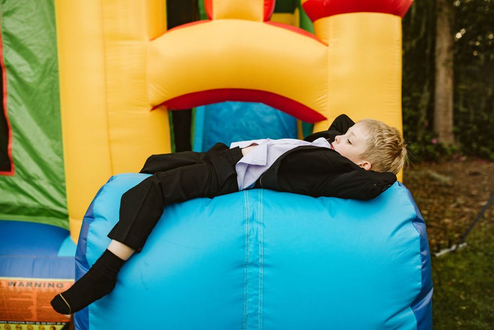 Young boy in formal suit relaxing on colorful inflatable bounce house at outdoor party.