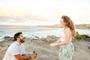 Woman reacting with joy as man proposes on one knee at scenic Hawaiian coastal overlook.