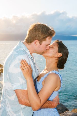 Couple sharing a romantic kiss by the ocean at sunset in matching blue Hawaiian outfits.