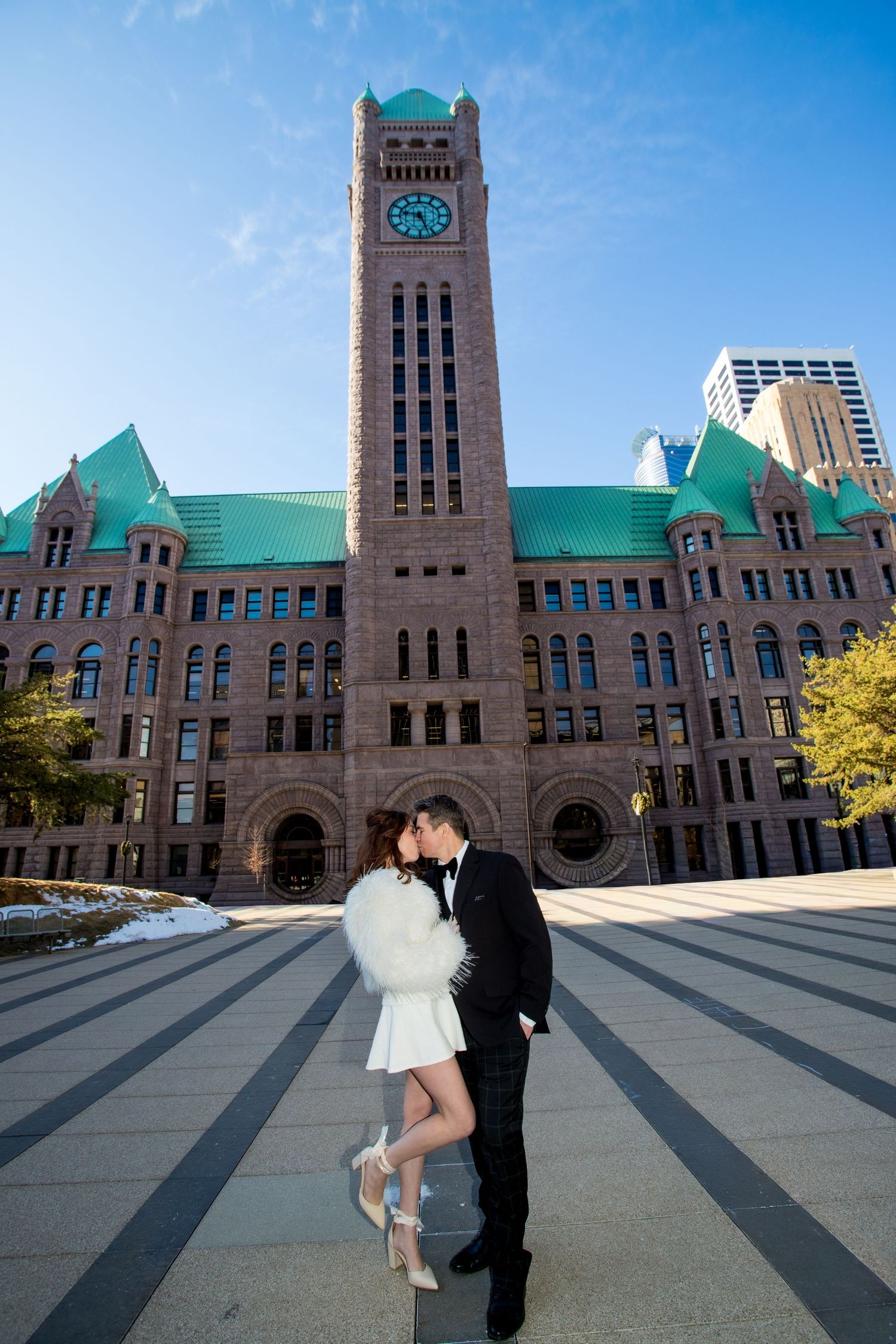 Couple poses in formal attire before Minneapolis City Hall clock tower on a sunny winter day.