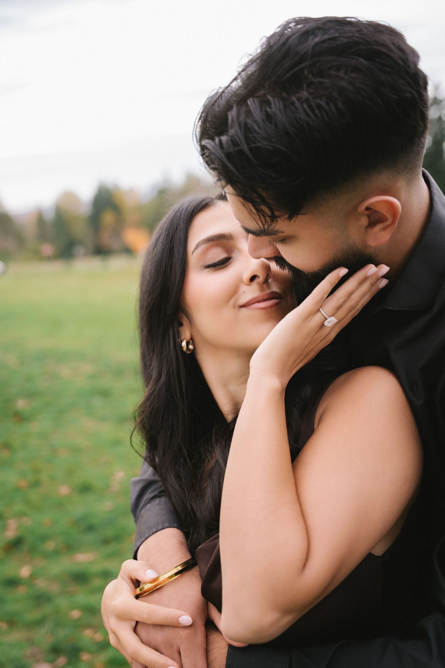 Couple in black outfits sharing a romantic embrace outdoors, woman displaying engagement ring, autumn park background.