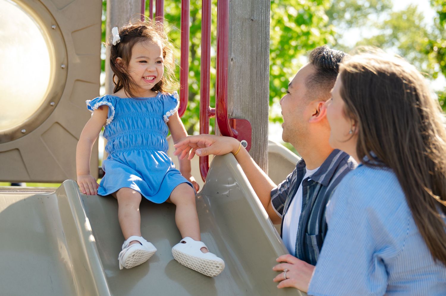 A young girl in a blue dress sits on a slide, smiling at two adults beside her on a sunny day during a family photoshoot in Tarrytown, NY.