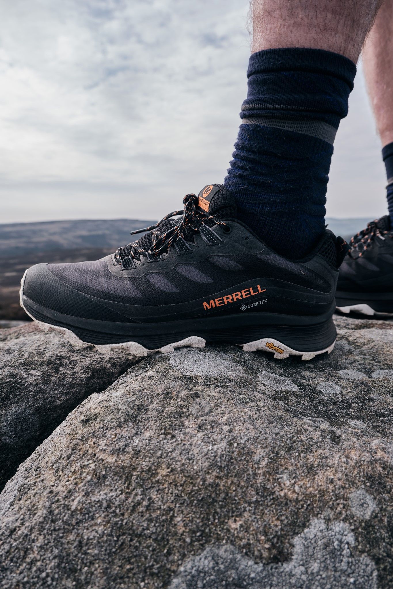 Black Merrell Gore-Tex hiking shoes on rocky terrain with dark socks, set against a cloudy sky backdrop.