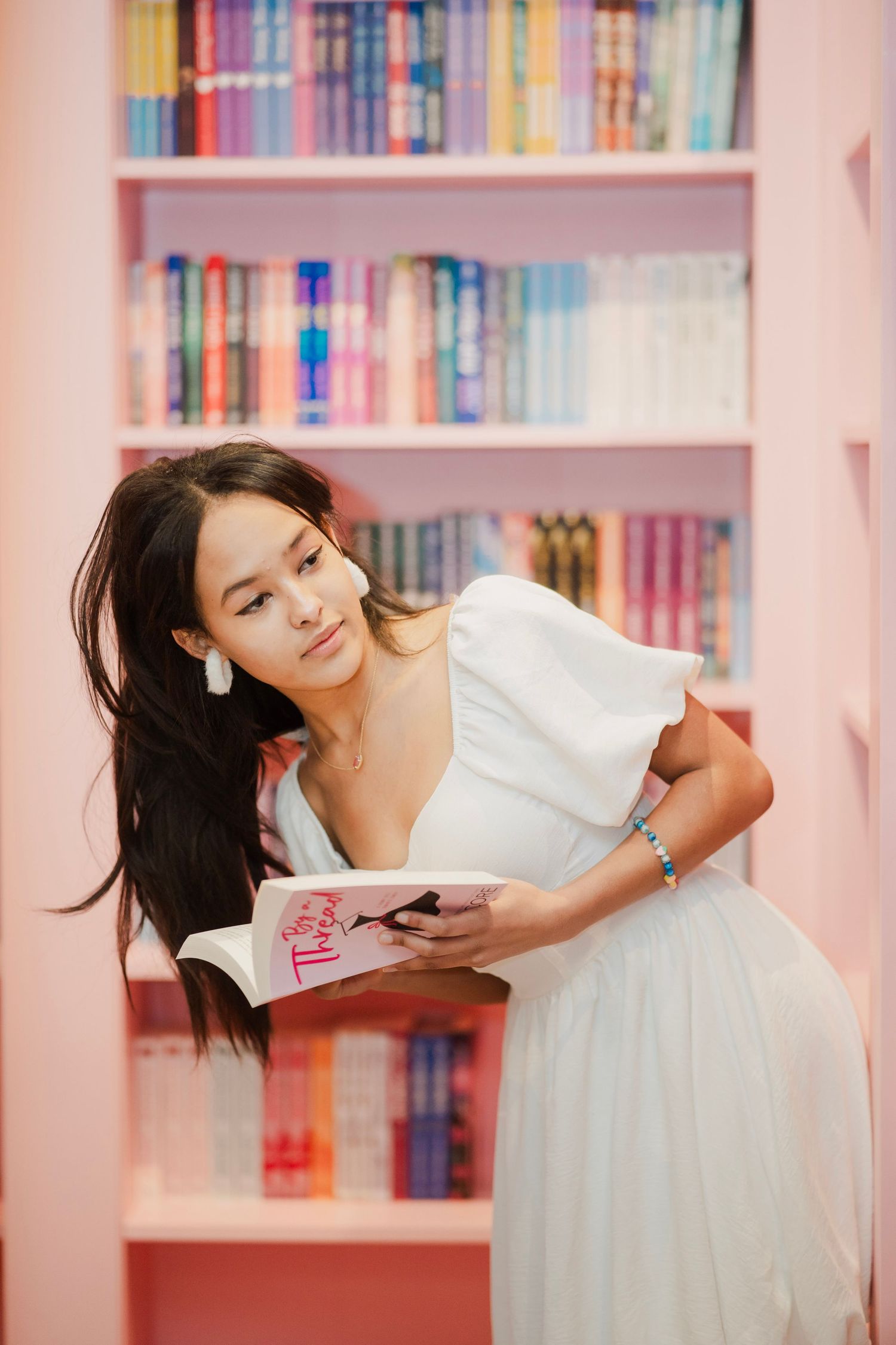 Young woman in white dress holds an open book while posing in front of colorful pink bookshelves.